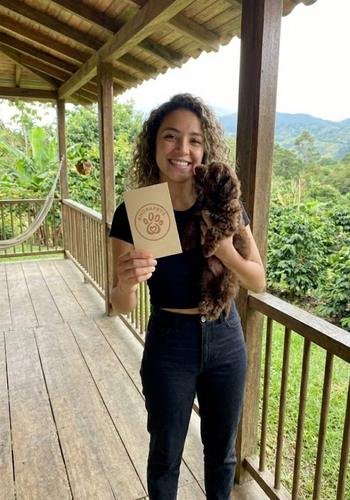 Chica joven recibiendo un cachorro Maltipoo de color café chocolate intenso en un parque con césped y árboles al fondo.